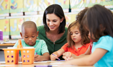 A teacher sits with three children who are drawing with crayons