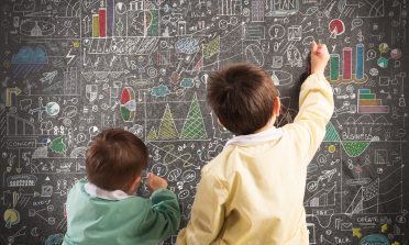 Two children write on a chalkboard that is full of charts and diagrams