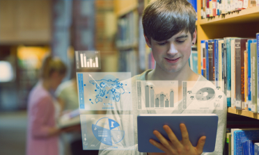 A boy is in a library with books on shelves behind him. He is looking at a screen which shows charts and graphs