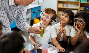 A teacher shows a test tube full of green liquid to three children wearing eye protection and smiling