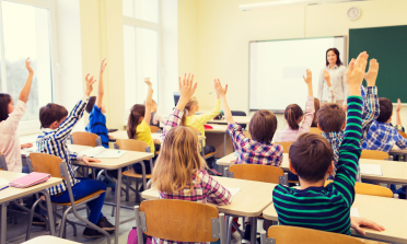 A classroom with children sitting at their desks, with their hands in the air.