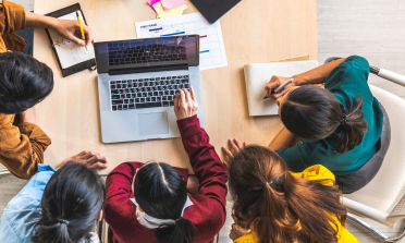 A group of people sit around a laptop at a desk, with notebooks and pens