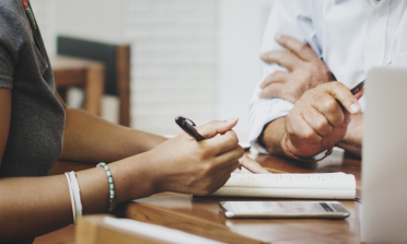 Two people's hands are holding pens over notepads on a desk with a laptop