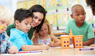 A teacher smiles as they help three children with their work at a desk