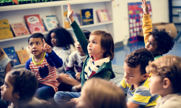 A group of young children sit on the floor. Three of them have their hands in the air.