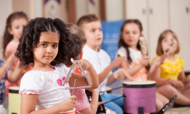 A girl plays a triangle and children in the background play drums and other instruments