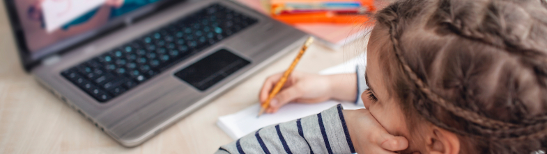 A girl writes on a notepad while watching a teacher on a laptop screen