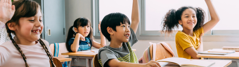 Four children sit smiling at desks in a classroom, with their hands raised in the air