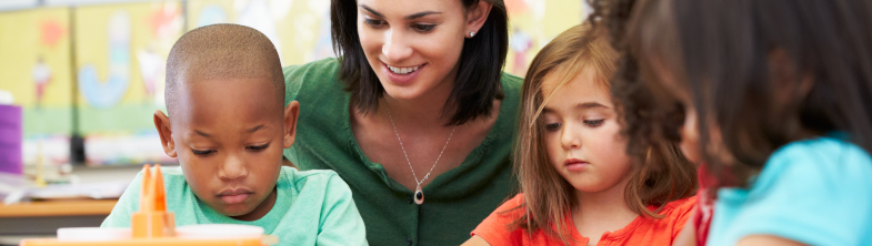 A teacher sits with three children who are drawing with crayons