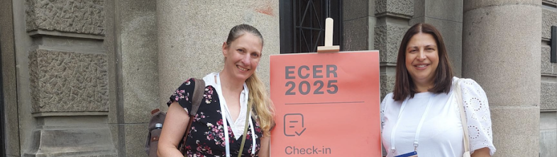Two people stand either side of a sign that reads 'ECER 2025 check in'
