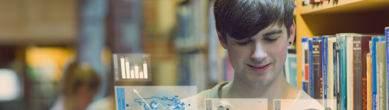 A boy is in a library with books on shelves behind him. He is looking at a screen which shows charts and graphs