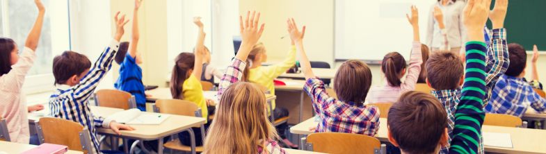 A classroom with children sitting at their desks, with their hands in the air.