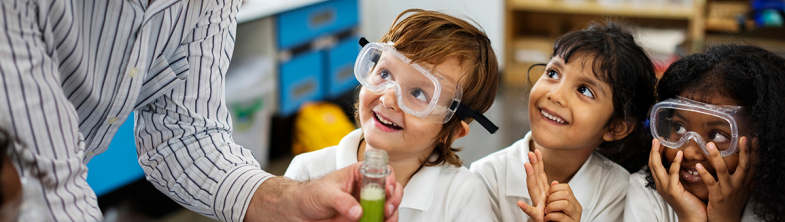 Three children wearing safety goggles smile at their teacher who is holding a test tube with a green liquid inside