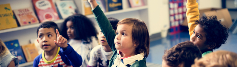 A group of young children sit on the floor. Three of them have their hands in the air.