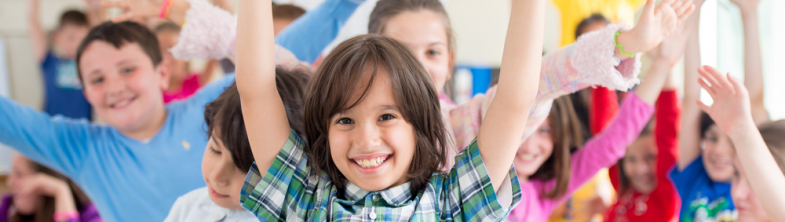 chidren in a classroon smiling and holding their hands high