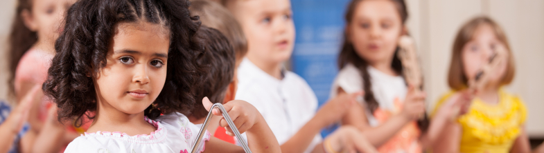 A girl plays a triangle and children in the background play drums and other instruments