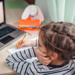 A girl writes on a notepad while watching a teacher on a laptop screen