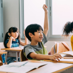 Four children sit smiling at desks in a classroom, with their hands raised in the air