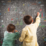 Two children write on a chalkboard that is full of charts and diagrams