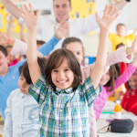 A group of children in a classroom wearing brightly coloured clothes and waving their arms in celebration