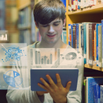 A boy is in a library with books on shelves behind him. He is looking at a screen which shows charts and graphs