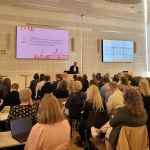 A person presenting at the front of a room full of audience members sitting on chairs