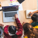 A group of people sit around a laptop at a desk, with notebooks and pens