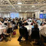 Participants at the bi-annual meeting sit around round tables and watch a presentation on a screen at the front of the room