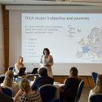 A person presenting at the front of a room full of audience members sitting on chairs. The screen behind the speaker says 'TCCA cluster 3 objective and countries'