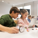 Two children look at a computer screen, one holding a magnifying glass