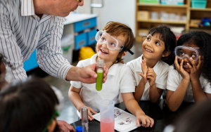 A teacher shows a test tube full of green liquid to three children wearing eye protection and smiling