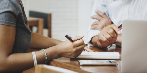 Two people's hands are holding pens as they take notes on a notepad