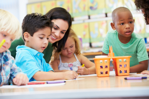 A teacher smiles as they help three children with their work at a desk