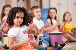 A girl plays a triangle and children in the background play drums and other instruments